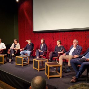 Syv paneldeltagere sitter på stoler på en scene.