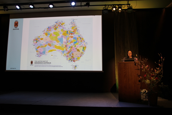 En kvinne st&aring;r bak en talerstol p&aring; en m&oslash;rk scene. Ved siden av henne viser en stor skjerm et kart over Australia.