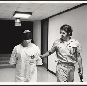 A prison guard leads a blindfolded prisoner through a corridor