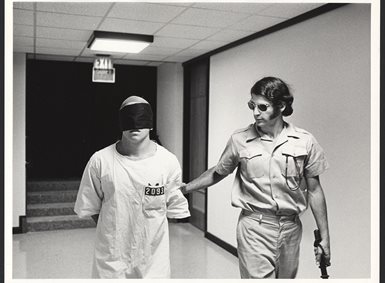 A prison guard leads a blindfolded prisoner through a corridor