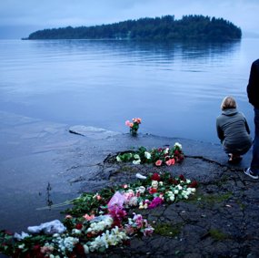 Two people stand on the shore by all the flowers laid down. They look over at Utøya.