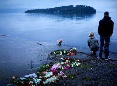 Two people stand on the shore by all the flowers laid down. They look over at Utøya.