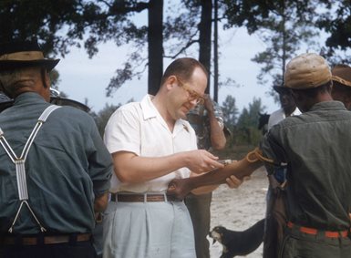 A Tuskegee Project researcher, a white man, takes a blood sample from a colored man outdoors.