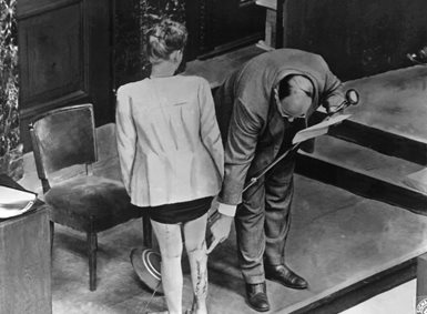 En mann i dress peker på lange arr på baksiden av leggen til en kvinne. Bildet er i sort-hvitt.