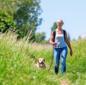 En voksen kvinne går tur med en hund over en grønn eng.
