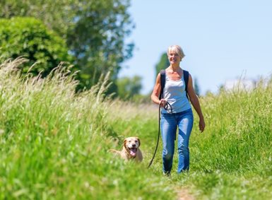 En voksen kvinne går tur med en hund over en grønn eng.