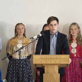 Oskar Hellesø-Knutsen, en ung mann i dress, står bak en talerstol og snakker. Bak ham står fire kvinner i samiske festdrakter.