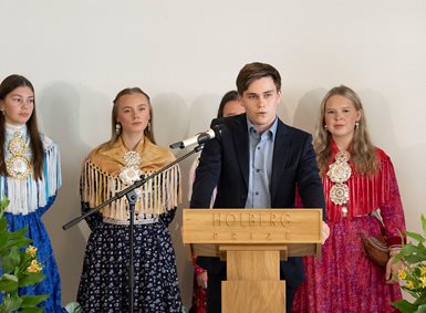 Oskar Hellesø-Knutsen, en ung mann i dress, står bak en talerstol og snakker. Bak ham står fire kvinner i samiske festdrakter.