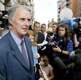 En godt voksen mann i grå dress står ute på gaten omringet av et stort pressekorps.