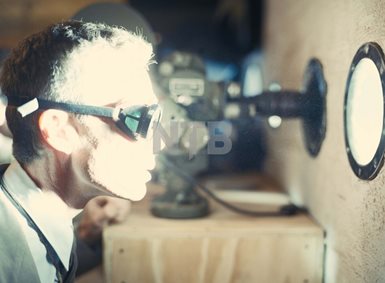 Cillian Murphy i rollen som Robert Oppenheimer ser med tykke beskyttelsesbriller inn gjennom et lite, rundt glassvindu. Det lyser sterkt fra vinduet på ansiktet hans.