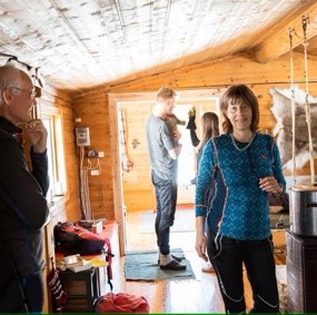 Marianne E. Lien står i et rom med trepanel, vedovn og reinsdyrskinn på veggen.
