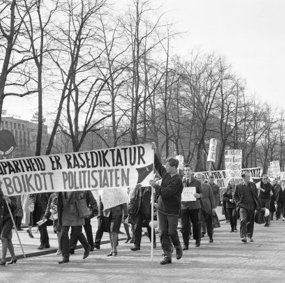 Mennesker i demonstrasjonstog bak fane med påskriften "Apartheid er rasediktatur - Boikott politistaten"